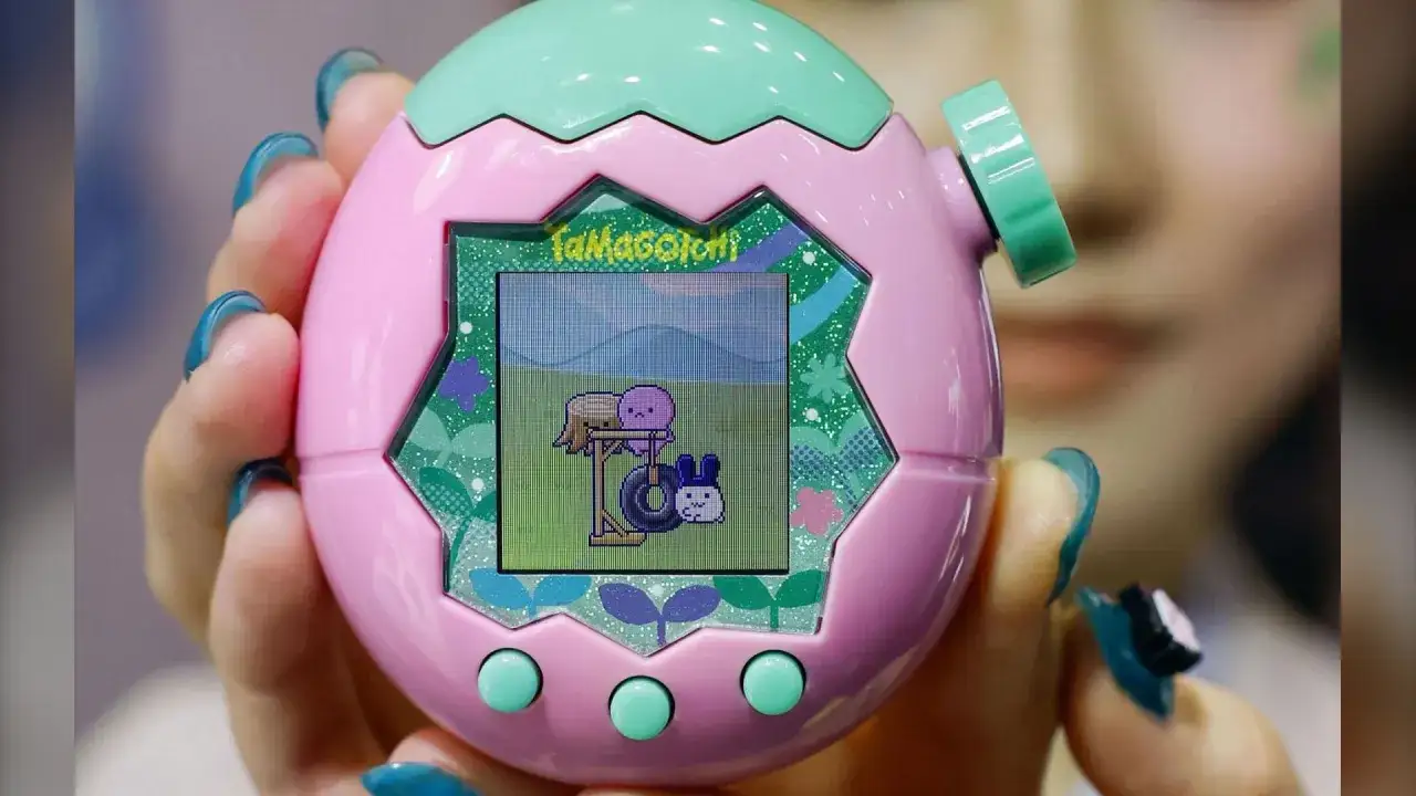 Różowe Tamagotchi z zielonymi przyciskami i ekranem, na którym widać wirtualne zwierzątko.